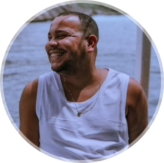 Foto de Mayk Brito sorrindo, usando óculos e camisa preta, barba e fundo amarelo.